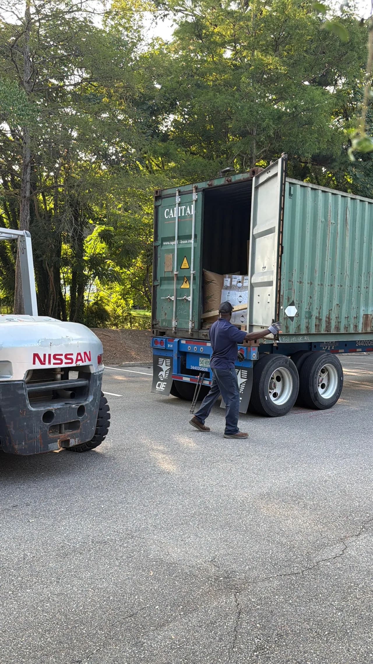 A worker standing next to an open shipping container on a trailer with a forklift parked nearby on the asphalt.