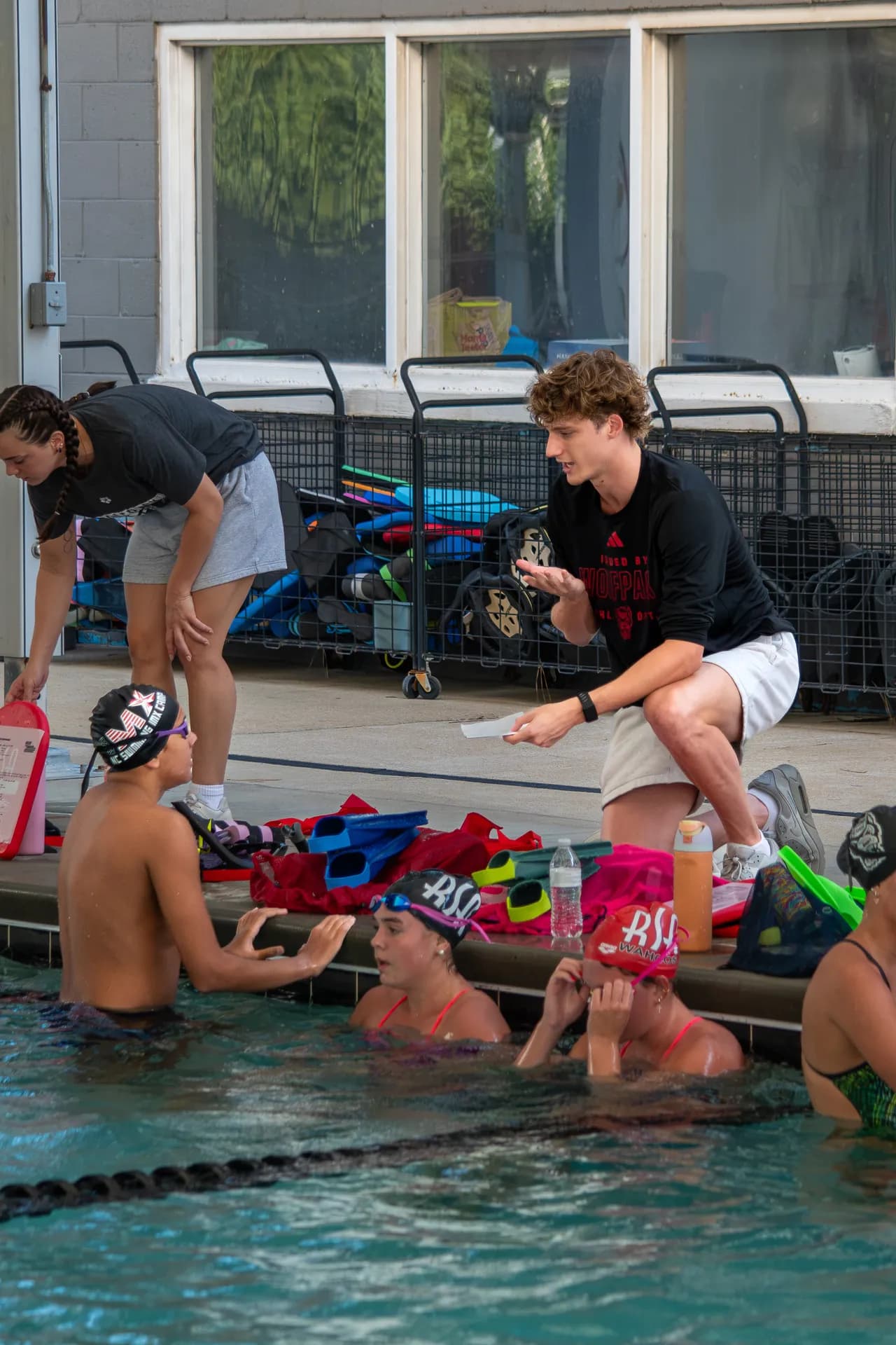 Wells Walker demonstrates a hand movement while speaking with swimmers gathered along the pool wall.