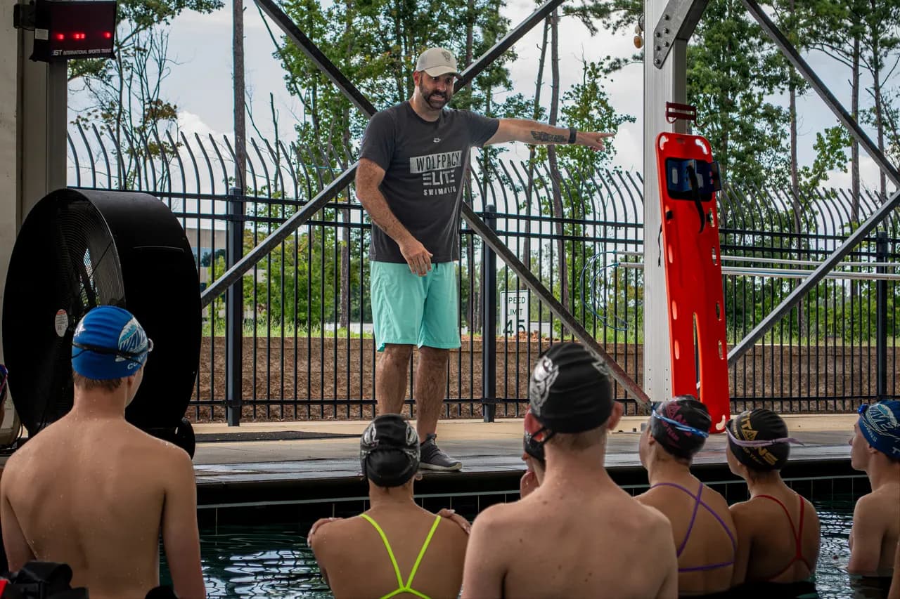 Braden Holloway points across the pool while explaining a drill to swimmers gathered along the pool wall.